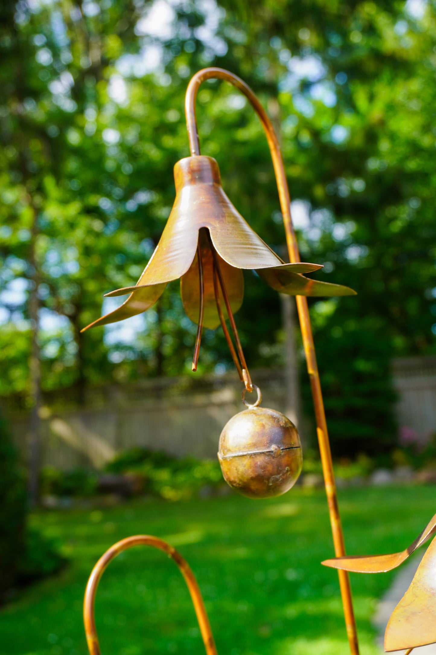 A bronze metal flower garden ornament with a bell-shaped flower head and a dangling bell, photographed outdoors in a garden with green grass and trees in the background.