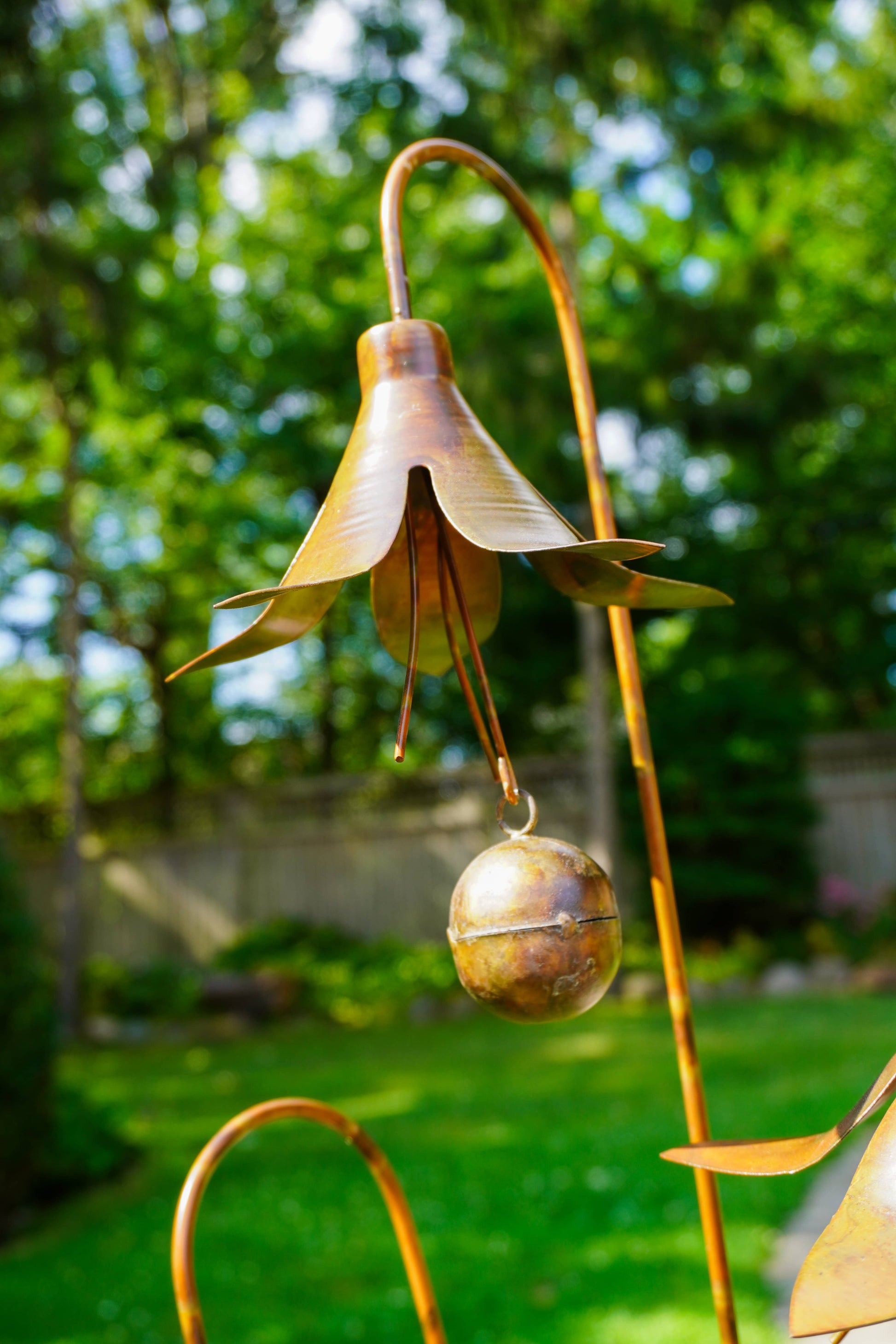 A bronze metal flower garden ornament with a bell-shaped flower head and a dangling bell, photographed outdoors in a garden with green grass and trees in the background.