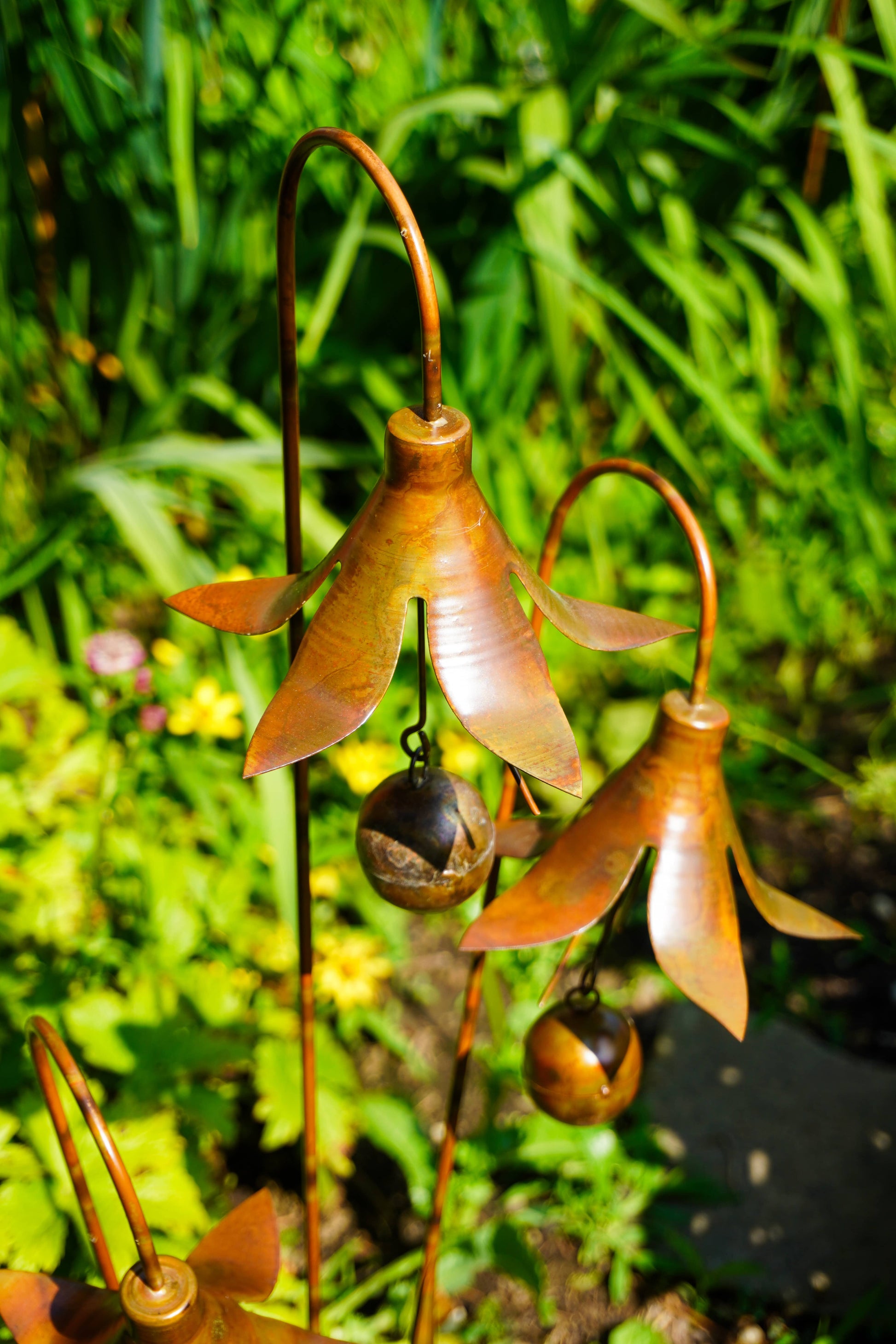 Metallic copper garden wind chimes shaped like bell-shaped flowers, with small hanging bells and curved wire stems, photographed outdoors in a garden with green foliage.
