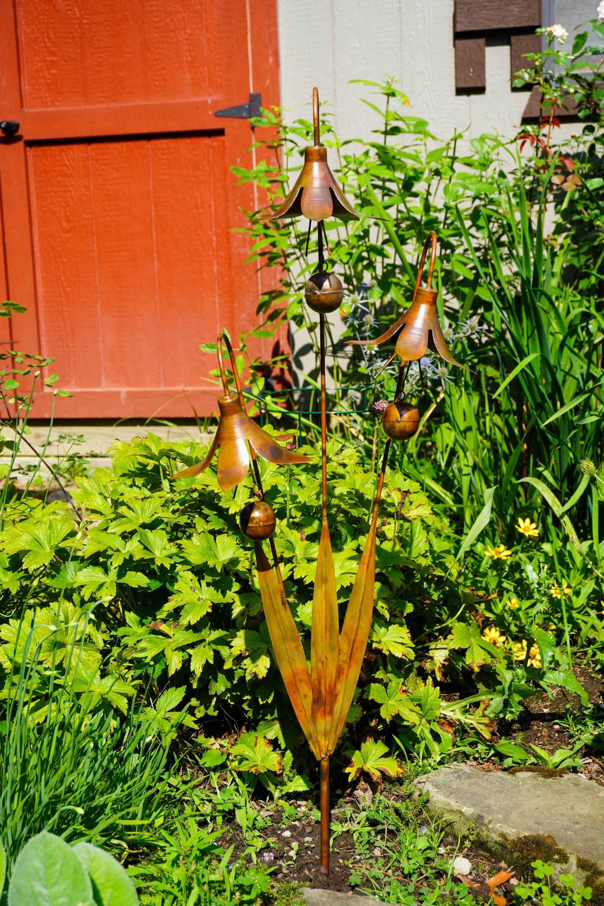 A copper-toned metal garden sculpture shaped like stylized flowers and leaves, placed in a garden bed with green plants and a red-orange door in the background.