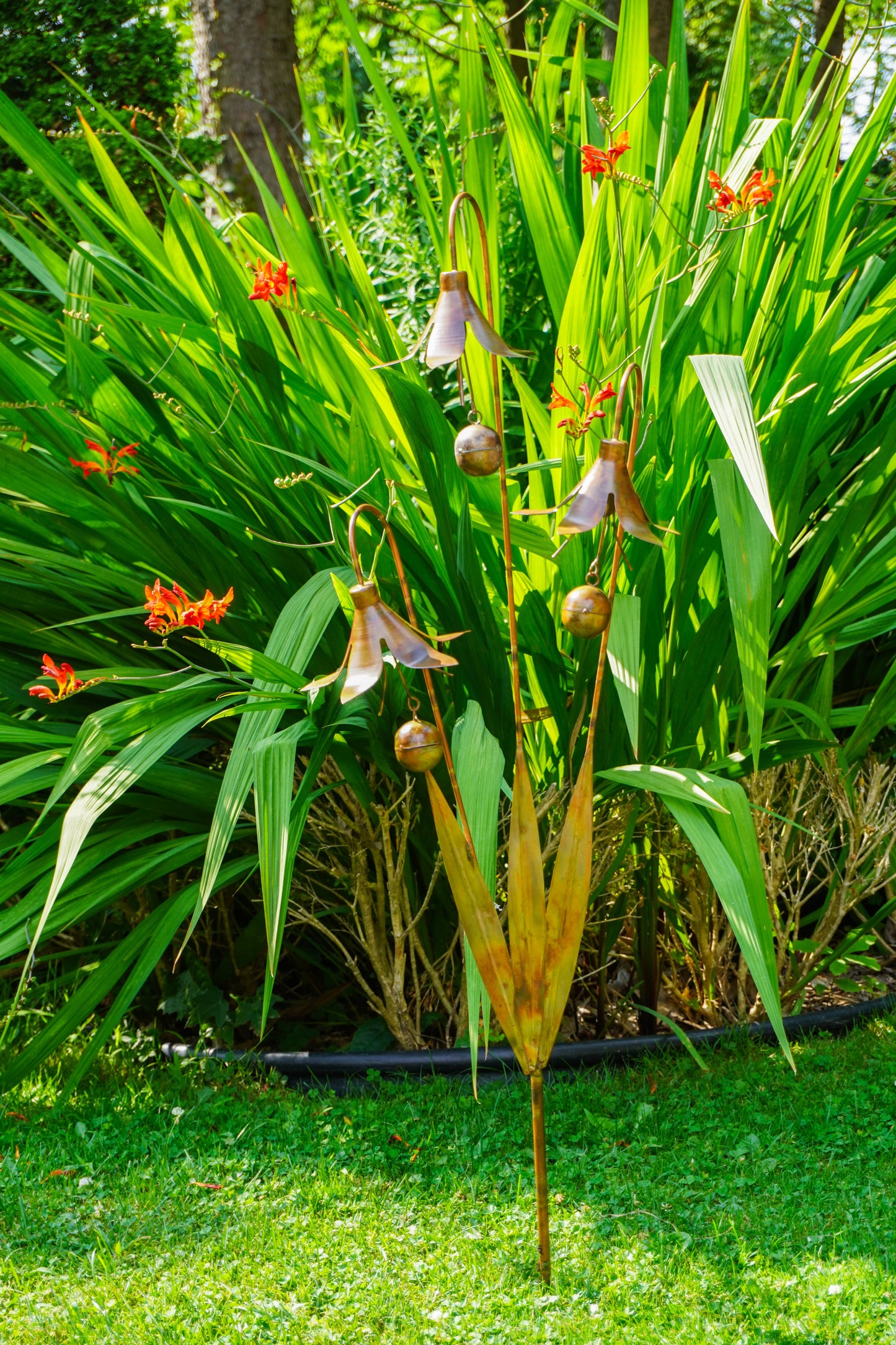 A garden sculpture depicting a stylized plant with hanging bell-shaped flowers and brown pods, set against a background of green tropical foliage and grass.