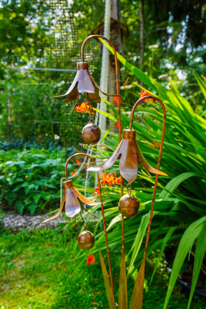 Copper metal garden sculpture shaped like hanging bellflowers with small hanging bells, photographed outdoors in a lush garden setting.