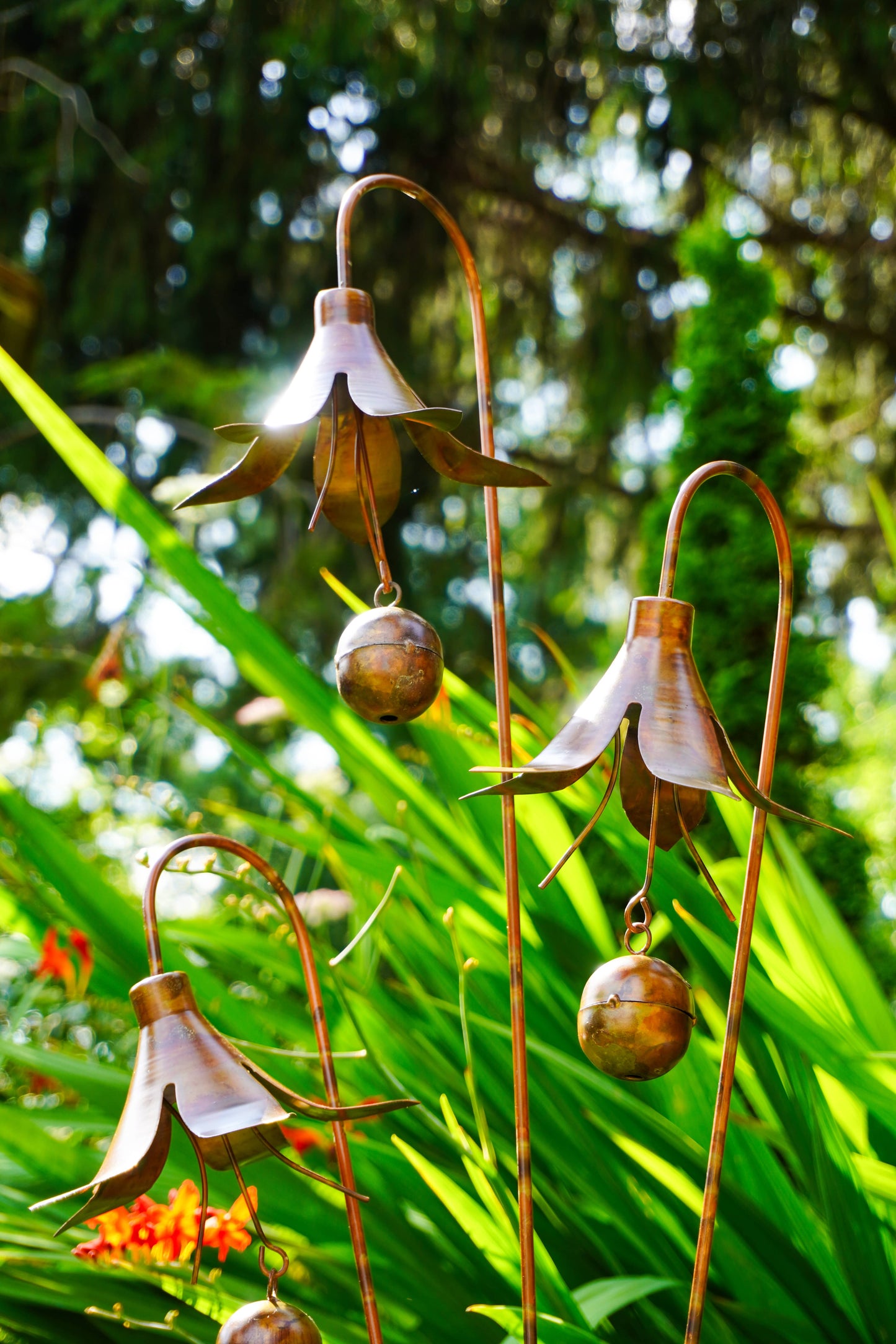 Metallic garden wind chimes shaped like bell-shaped flowers with small hanging bells, photographed outdoors among green plants and sunlight.