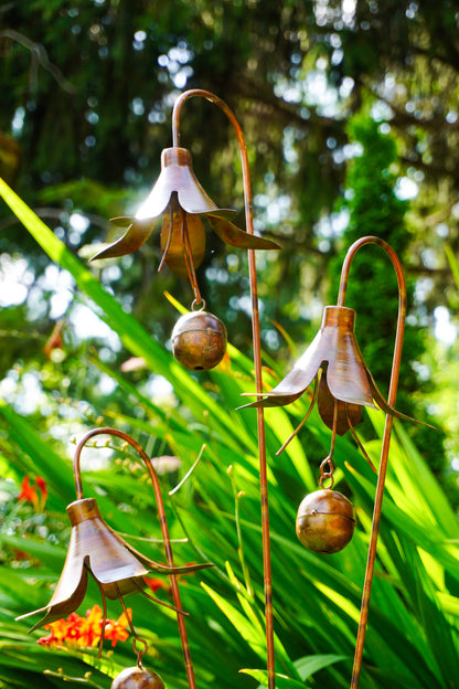 Metallic garden wind chimes shaped like bell-shaped flowers with small hanging bells, photographed outdoors among green plants and sunlight.