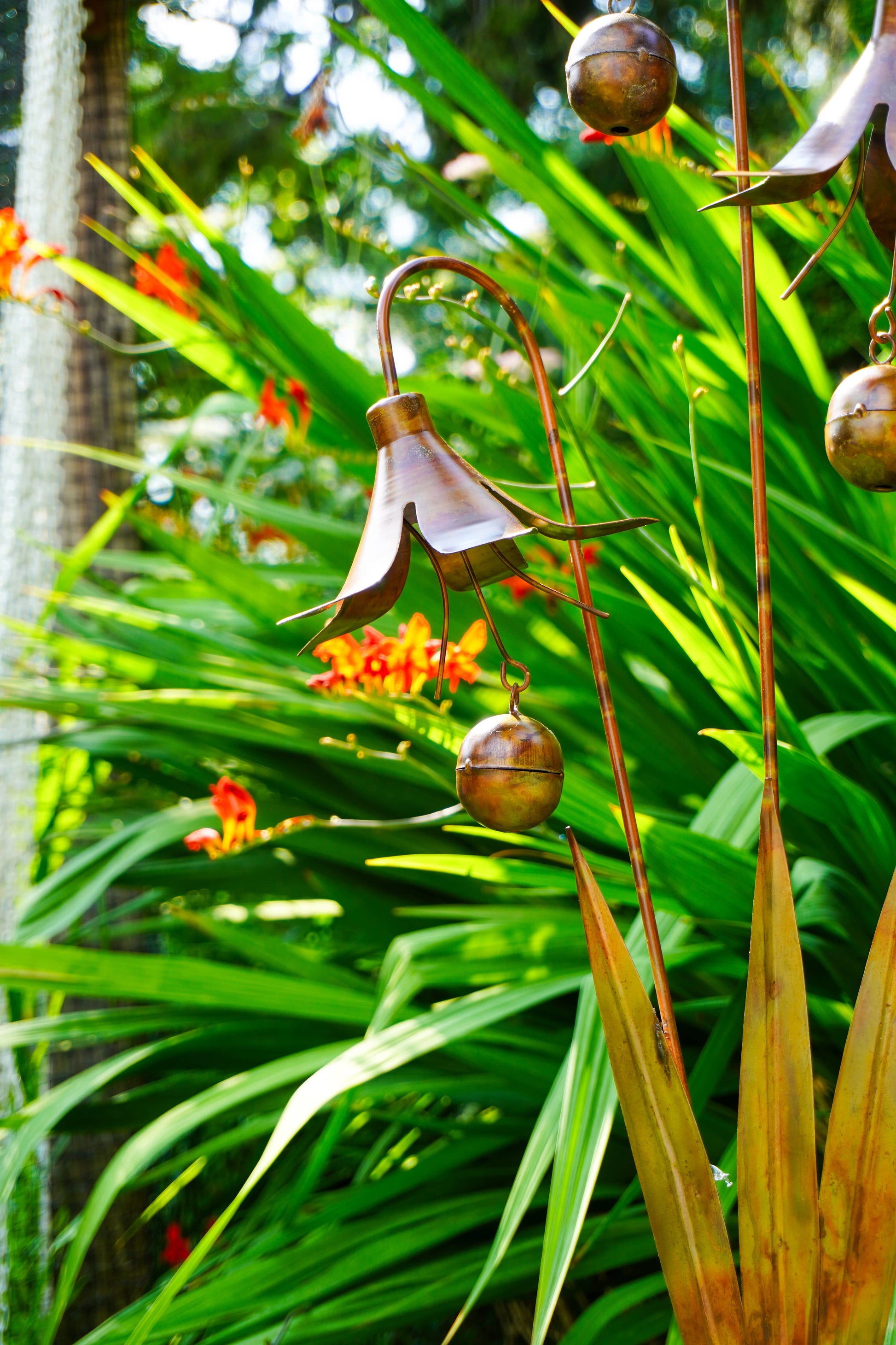 A copper metal garden sculpture shaped like a bell-shaped flower with hanging bells, displayed outdoors among green tropical plants and small orange flowers.