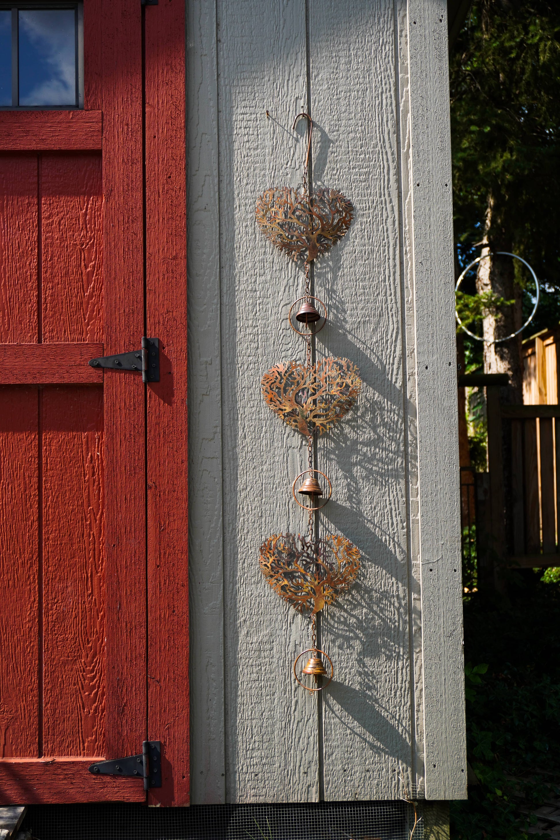 Three rustic metal heart-shaped wind chimes with small hanging bells are displayed vertically on a white textured wall next to a red wooden door.