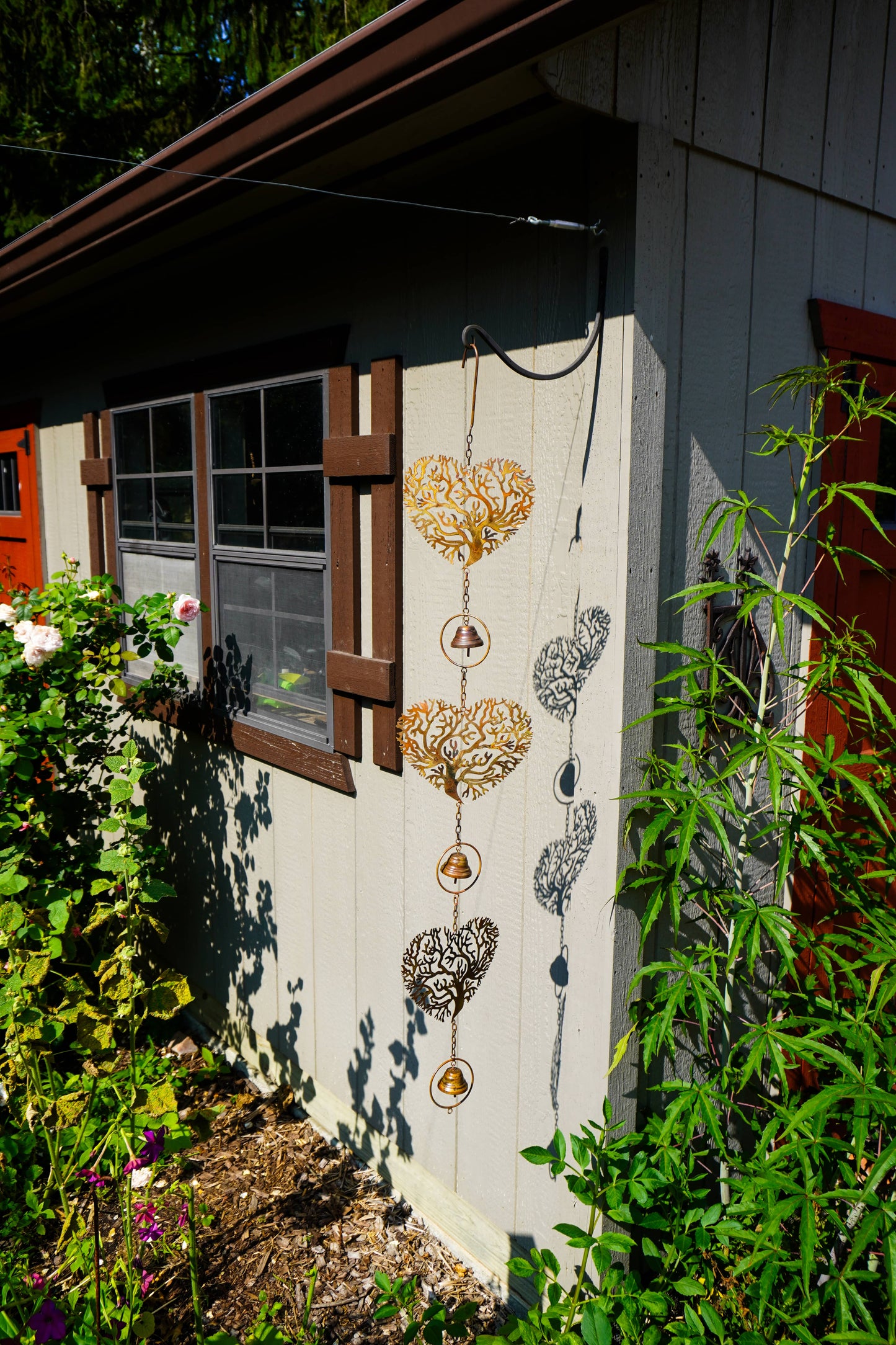 Three hanging heart-shaped metal garden wind chimes in gold, silver, and bronze finishes are displayed on the exterior wall of a house, casting shadows on the siding.