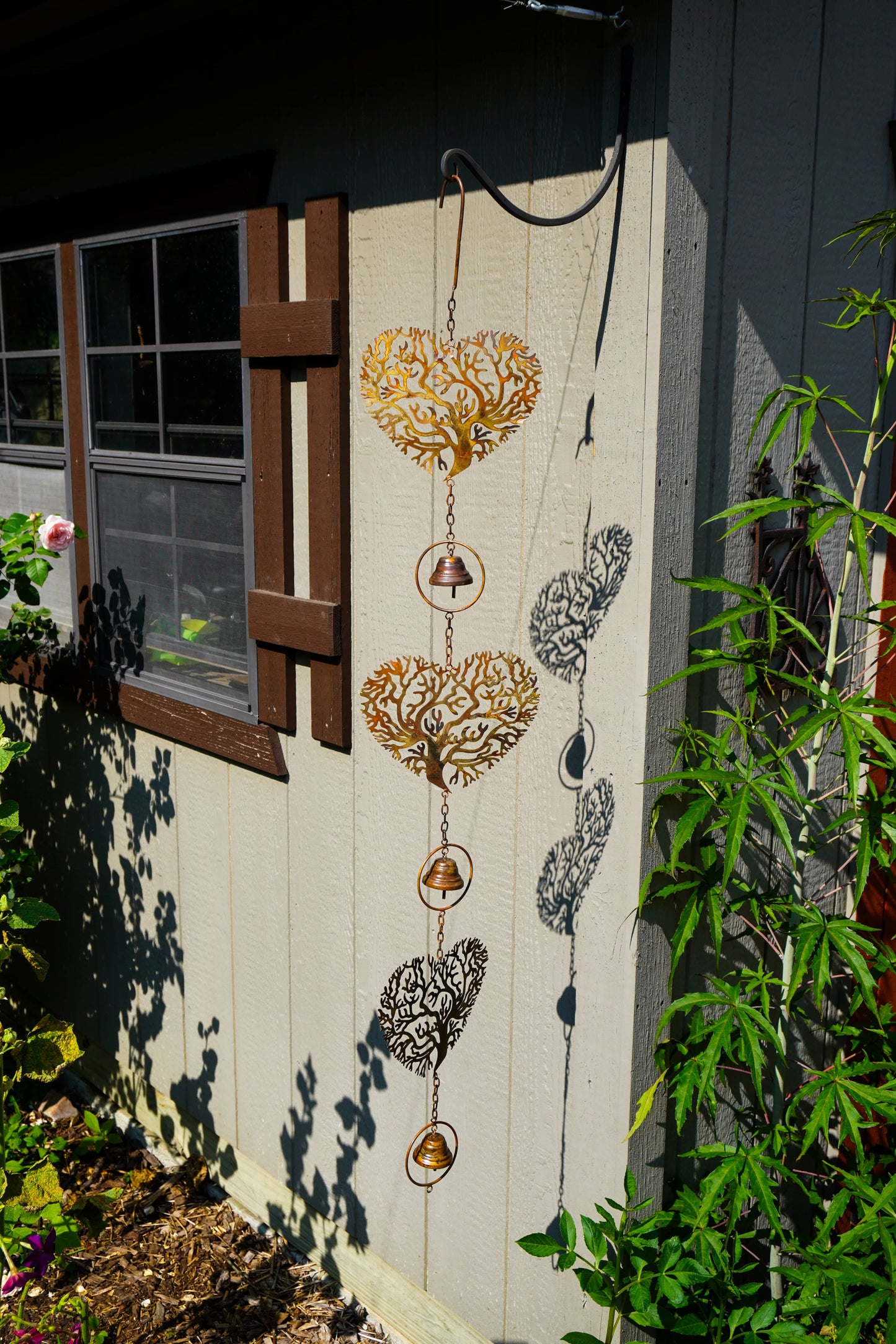 A decorative metal wind spinner hanging on a light-colored wooden fence in a garden setting. The spinner features three gold-toned heart-shaped windcatchers with intricate cutout patterns, connected by chains to small hanging bells and round windweights, with two additional black-and-white patterned heart windcatchers below.