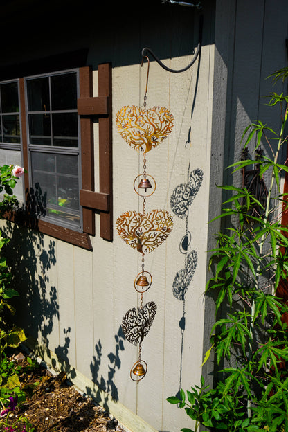 A decorative metal wind spinner hanging on a light-colored wooden fence in a garden setting. The spinner features three gold-toned heart-shaped windcatchers with intricate cutout patterns, connected by chains to small hanging bells and round windweights, with two additional black-and-white patterned heart windcatchers below.
