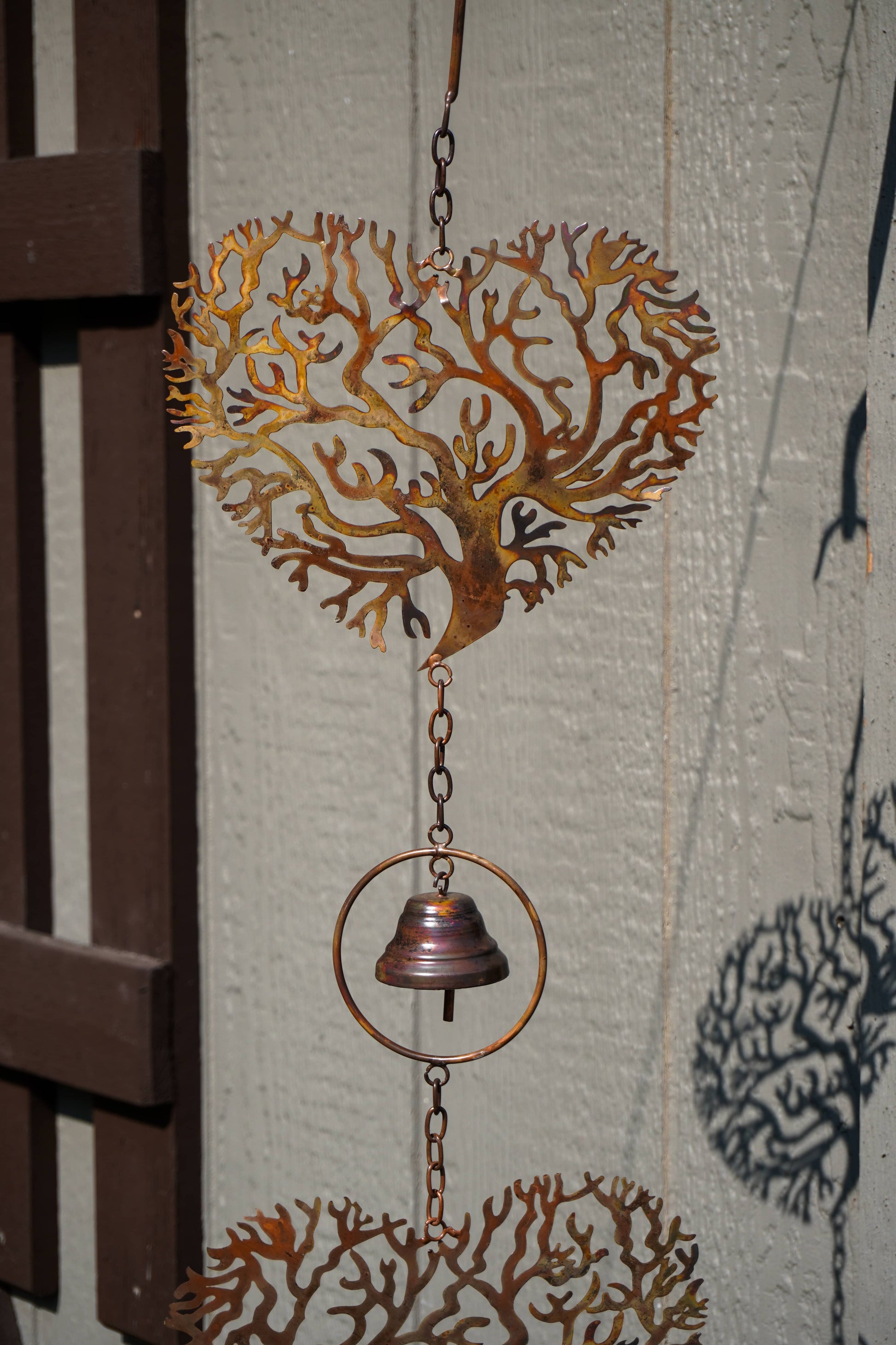 A rusted copper metal wind spinner featuring a heart-shaped coral tree design, a hanging bell, and a circular ring, shown outdoors against a textured wall.