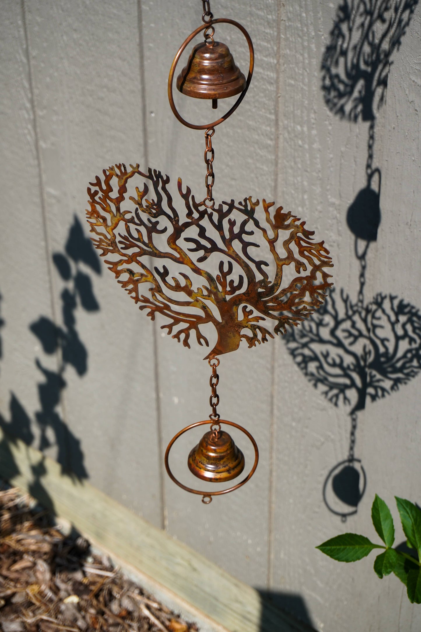 A copper-colored metal wind spinner hanging outdoors against a gray wood-paneled wall. The design features a heart-shaped tree silhouette in the center, flanked by two circular rings with small hanging bells at the top and bottom.