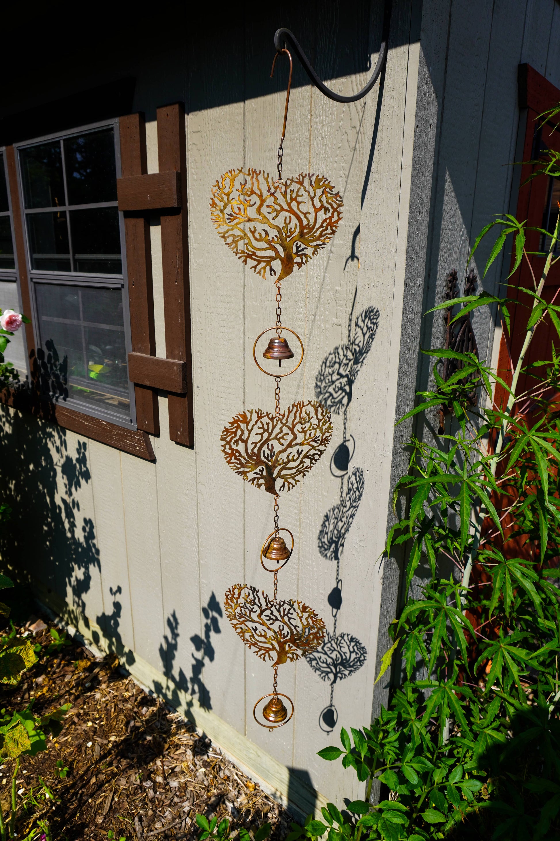 A decorative metal wind spinner hanging outdoors against a house wall, featuring three heart-shaped tree silhouettes with hanging bells and circular rings. The wind spinner is shown next to green plants and a window, with its shadow cast on the wall.