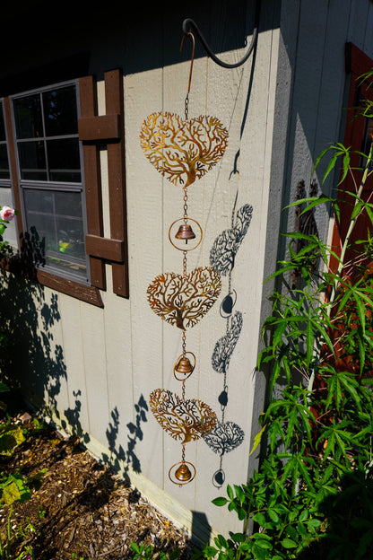 A decorative metal wind spinner hanging outdoors against a house wall, featuring three heart-shaped tree silhouettes with hanging bells and circular rings. The wind spinner is shown next to green plants and a window, with its shadow cast on the wall.