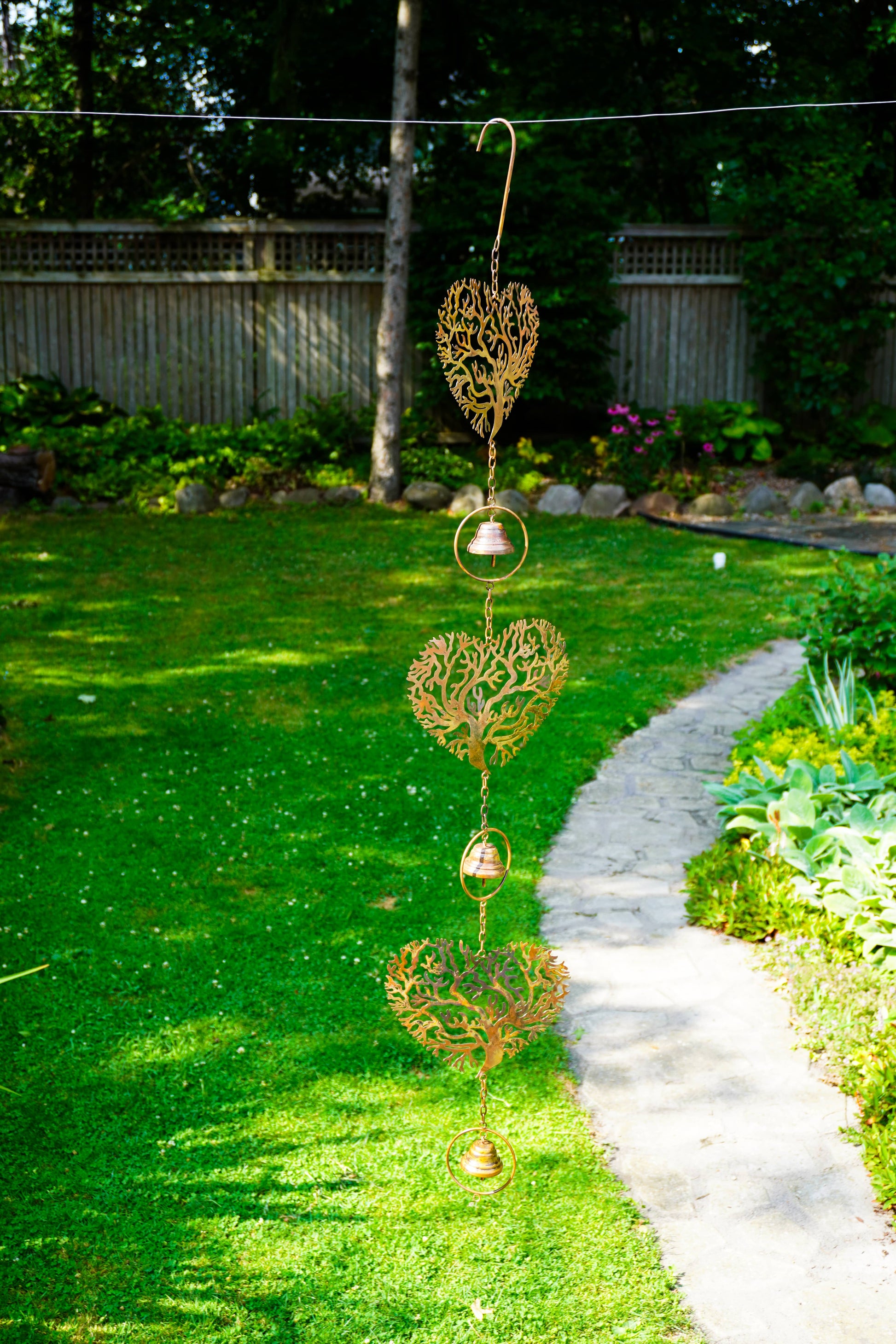 A hanging garden ornament with three ornate gold heart-shaped cutouts and small gold bells, suspended outdoors on a wire over a green lawn next to a stone path.