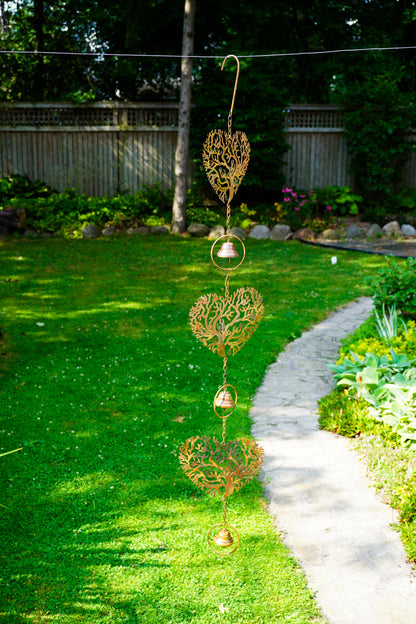 A hanging garden ornament with three ornate gold heart-shaped cutouts and small gold bells, suspended outdoors on a wire over a green lawn next to a stone path.