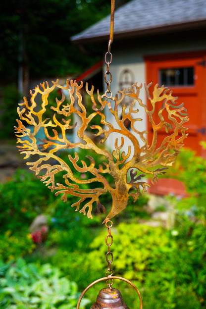 A rusted metal heart-shaped tree wind spinner hanging in a garden, with a small bronze bell at the bottom, set against a blurred background of green foliage and a red house.