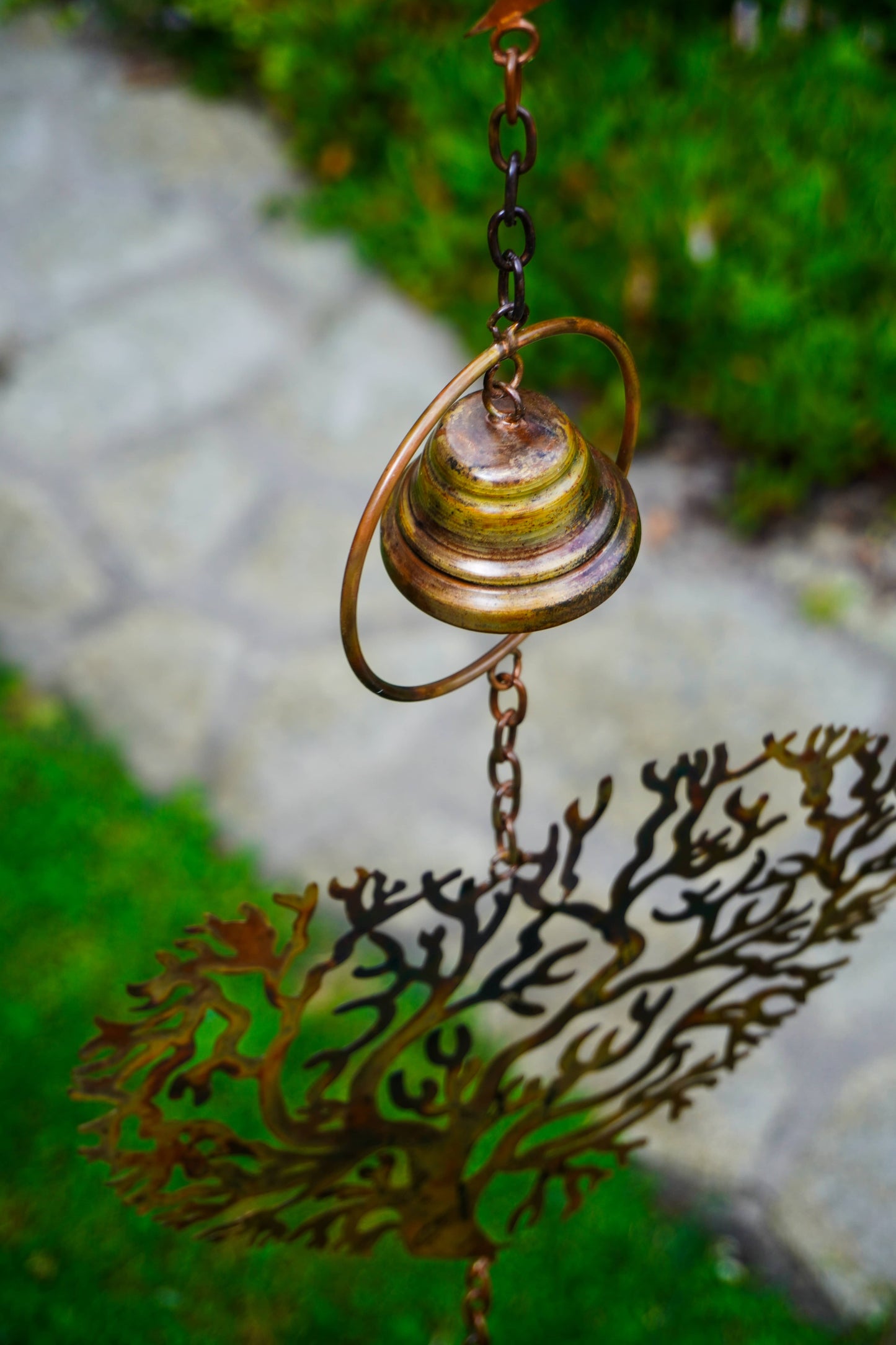 A bronze metal garden wind spinner hanging outdoors, featuring a central spinning ball on a chain above a coral-like leaf silhouette, with blurred green grass and a stone path in the background.