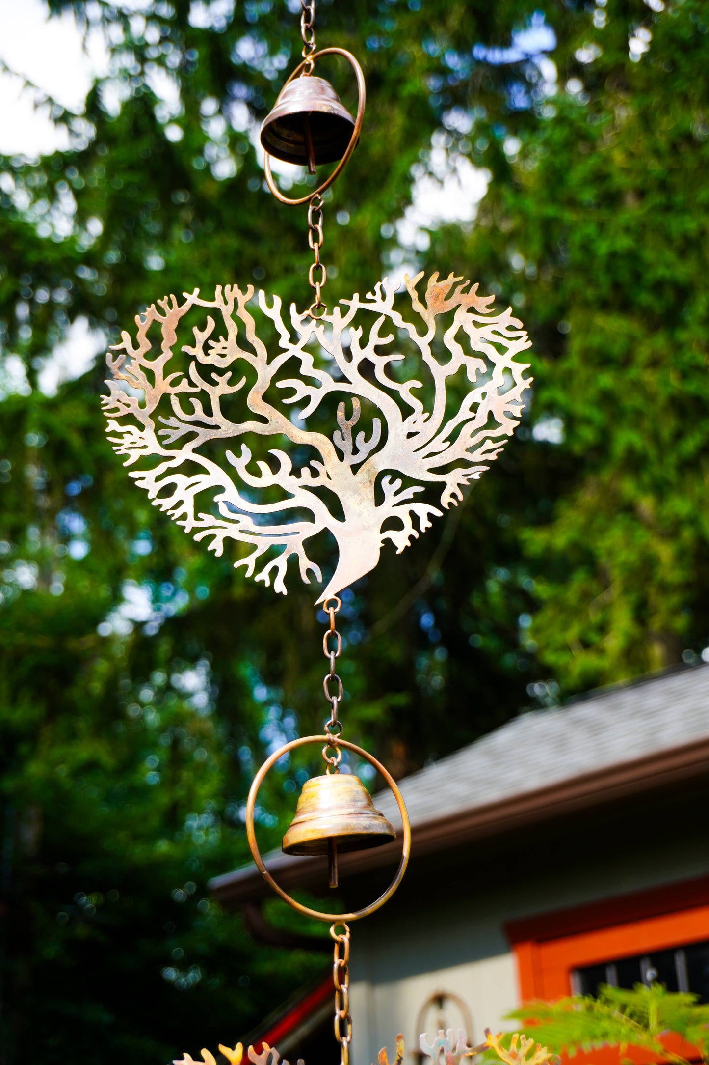 A decorative metal wind chime featuring a heart-shaped tree design with two hanging brass bells, photographed outdoors against a blurred background of green trees and a house.