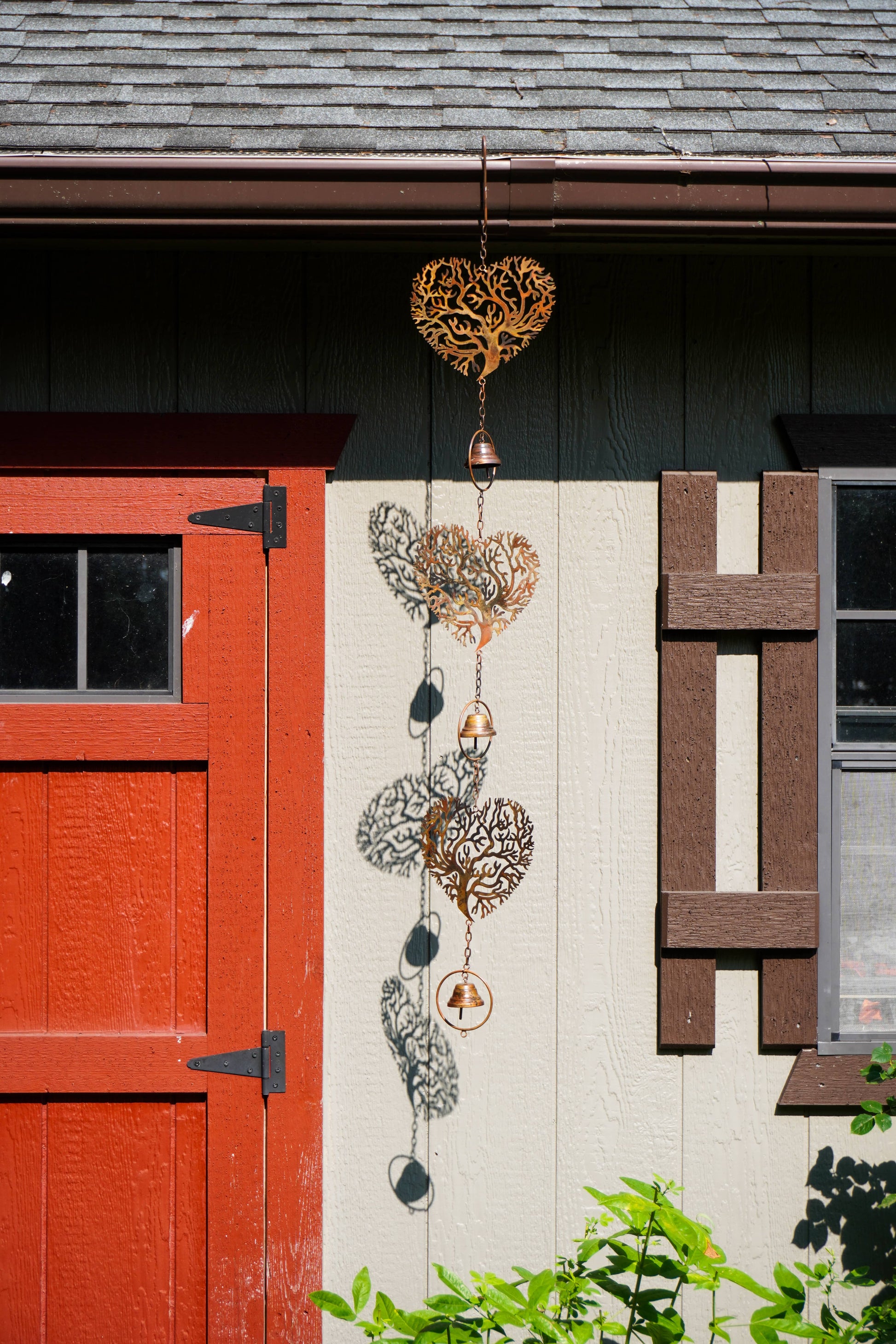 A decorative metal wind spinner with multiple heart-shaped cutout designs in gold, silver, and bronze tones, hanging outdoors against a house wall next to a red door.