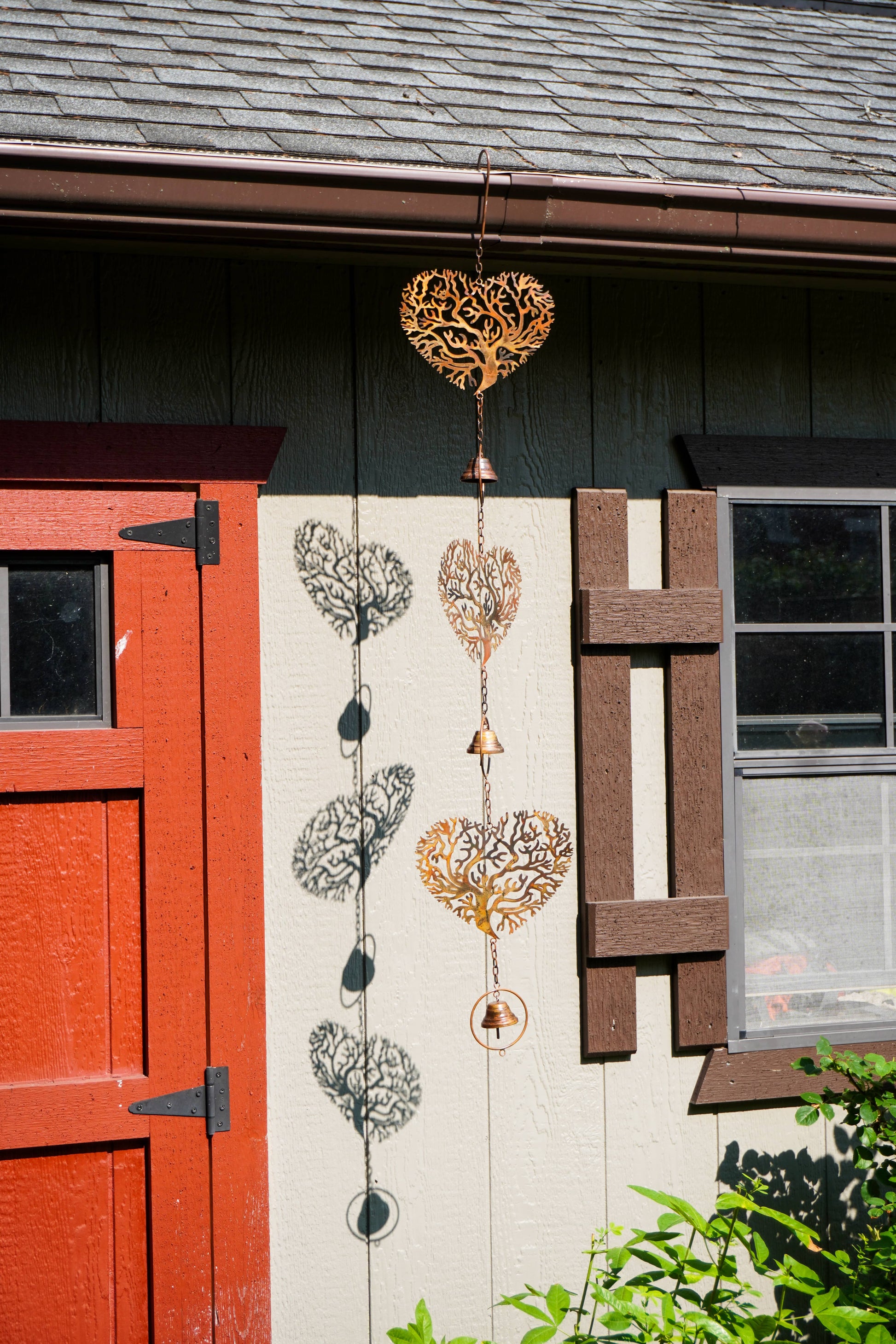 Two decorative metal wind spinner ornaments hanging from a house exterior. One ornament is gold and the other is black, both featuring tree-of-life heart motifs and hanging bells.