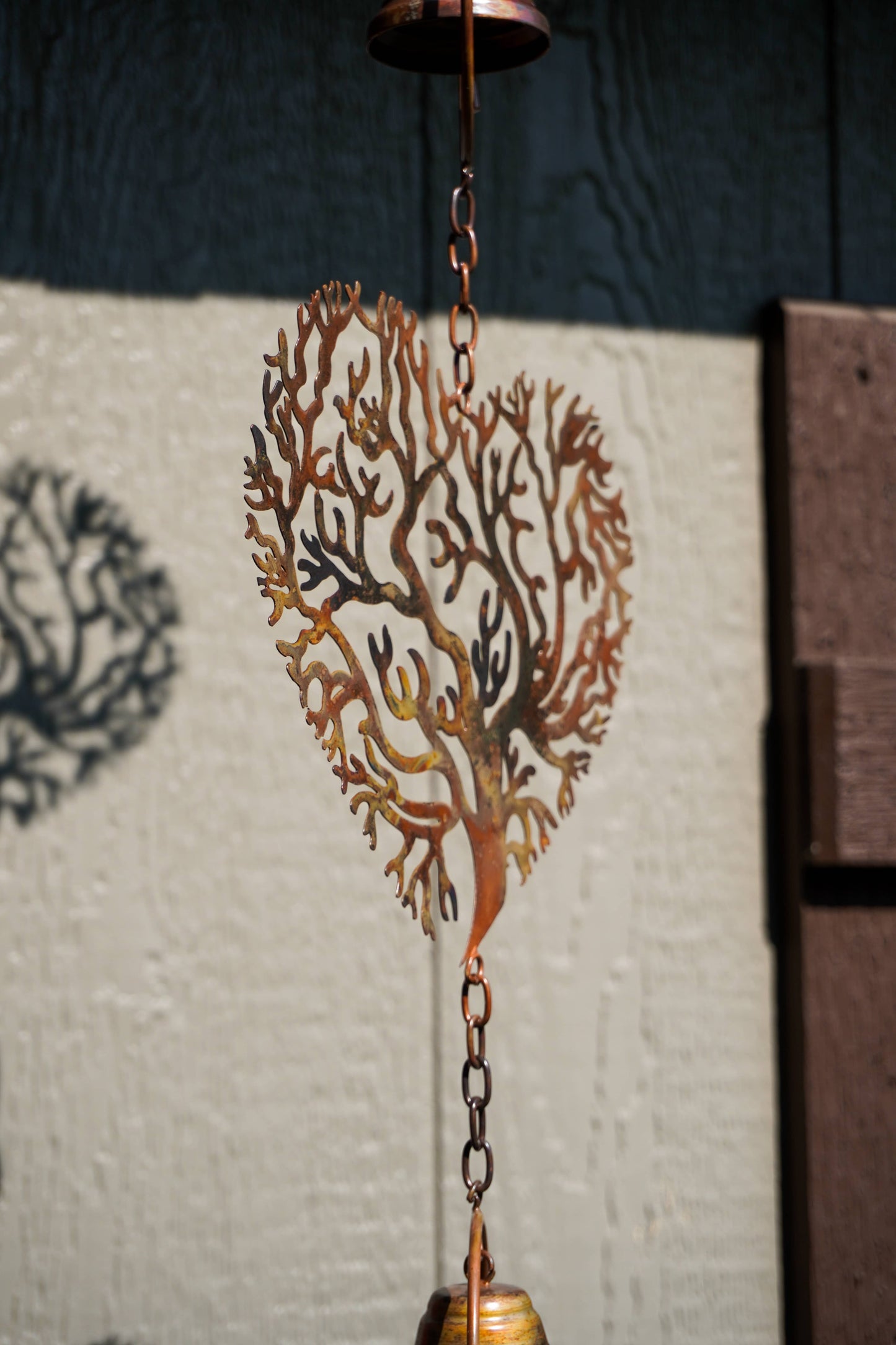 A decorative metal wind spinner shaped like a tree branch, hanging from chains with a small bell at the bottom, photographed outdoors against a textured wall.