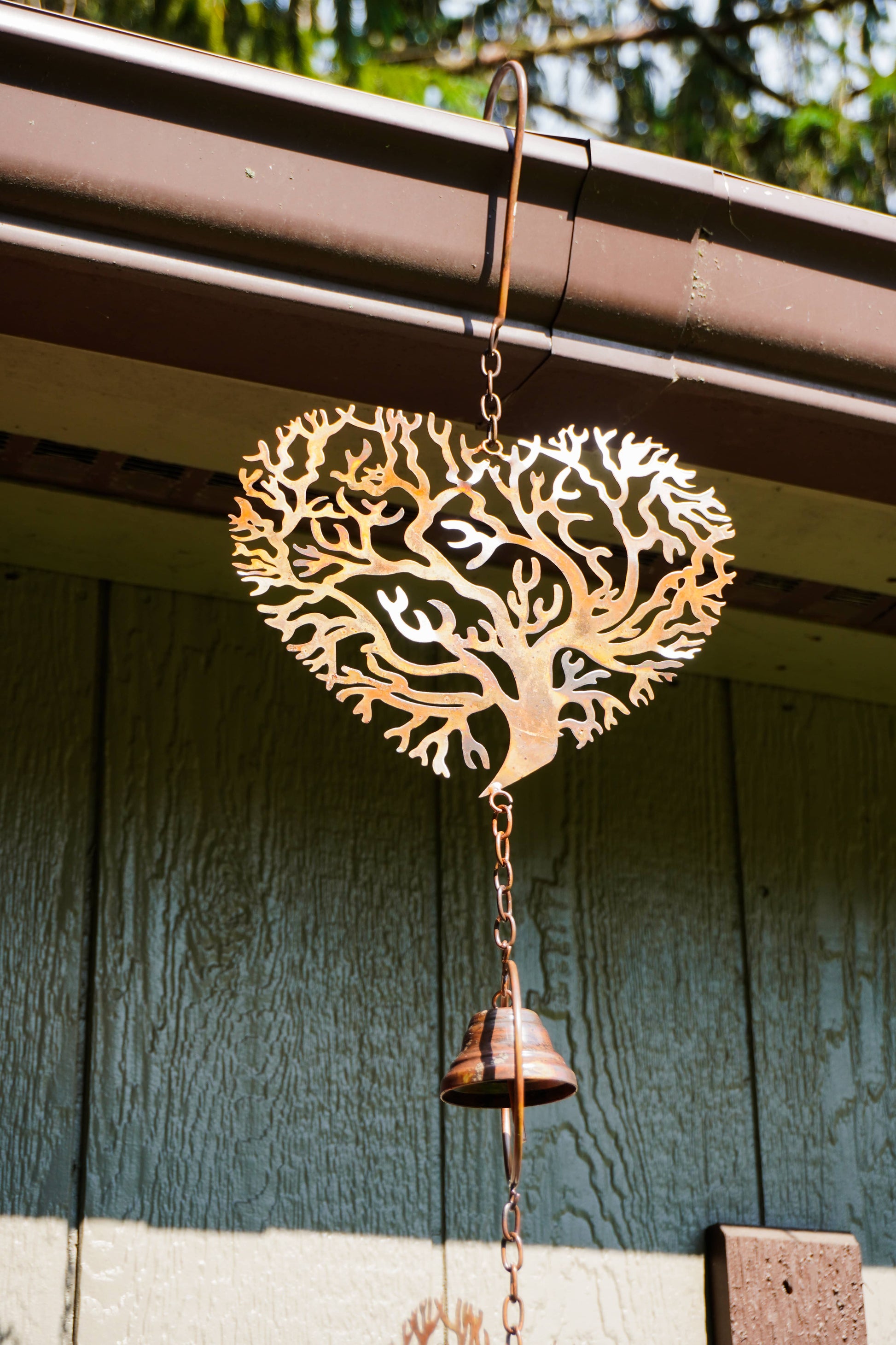 A copper-colored metal heart-shaped wind chime with a laser-cut tree design hangs from a brown gutter above a green wooden wall. A small hanging bell is suspended below the heart by a chain.