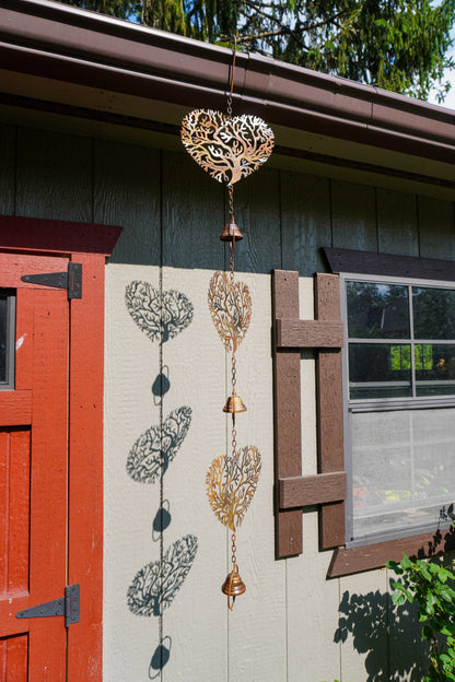 A decorative metal wind spinner with heart-shaped tree silhouettes and small hanging bells, shown outdoors attached near the eaves of a house next to a red door and window.