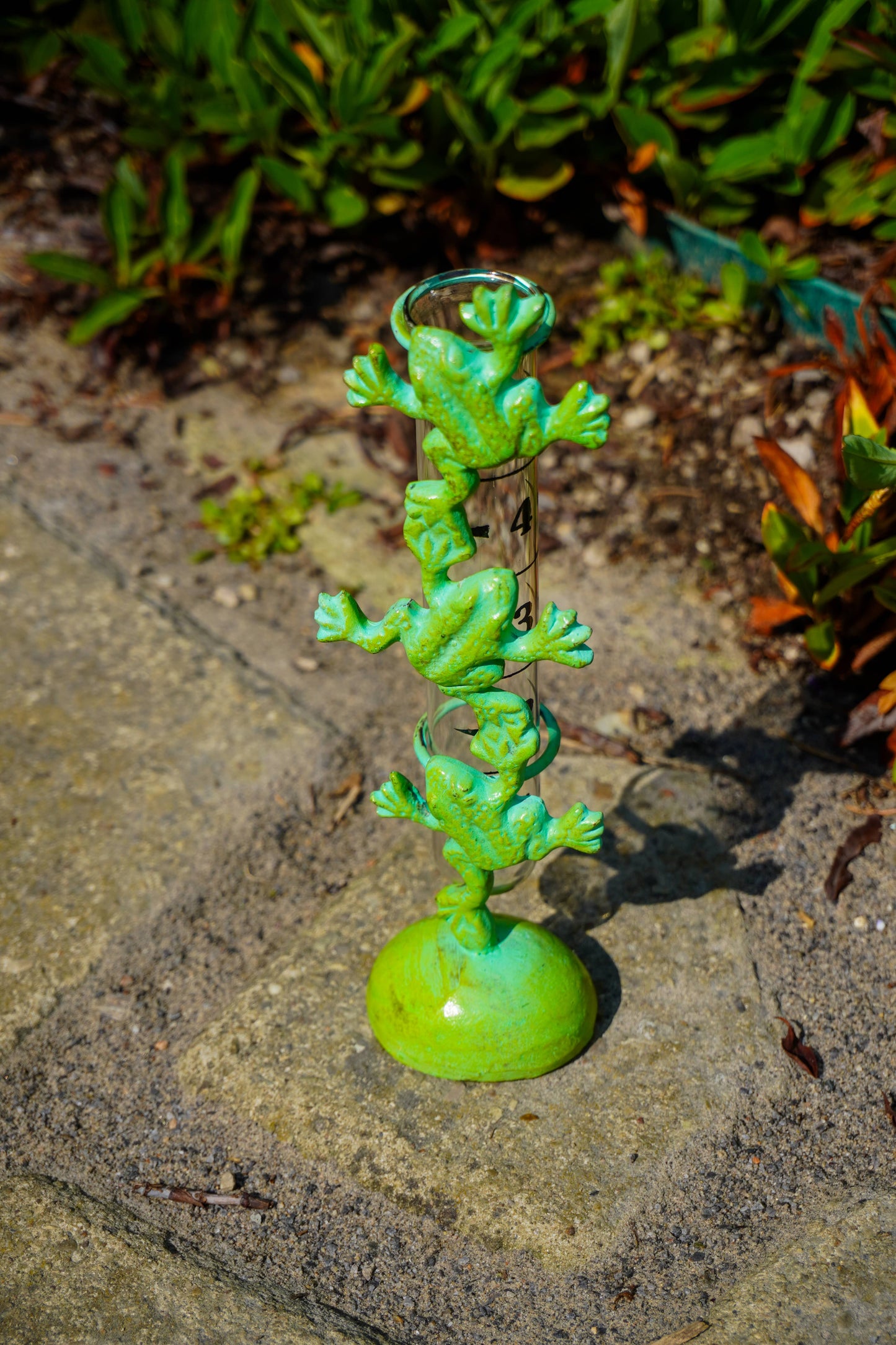 A green frog-themed metal garden stake with three stacked frogs and a hanging ring, photographed outdoors on a stone path with blurred plants in the background.