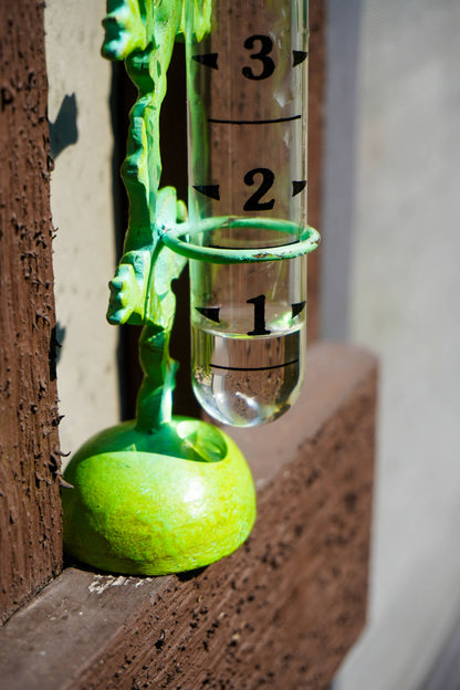 A close-up of a green decorative object with a clear cylindrical tube marked 1, 2, and 3, mounted against a textured brown surface.