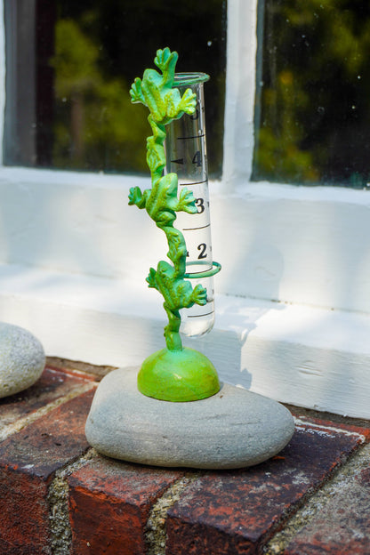 A green plant-shaped rain gauge with a clear measuring tube and gray stone base, set outdoors on a brick surface in front of a white window frame.