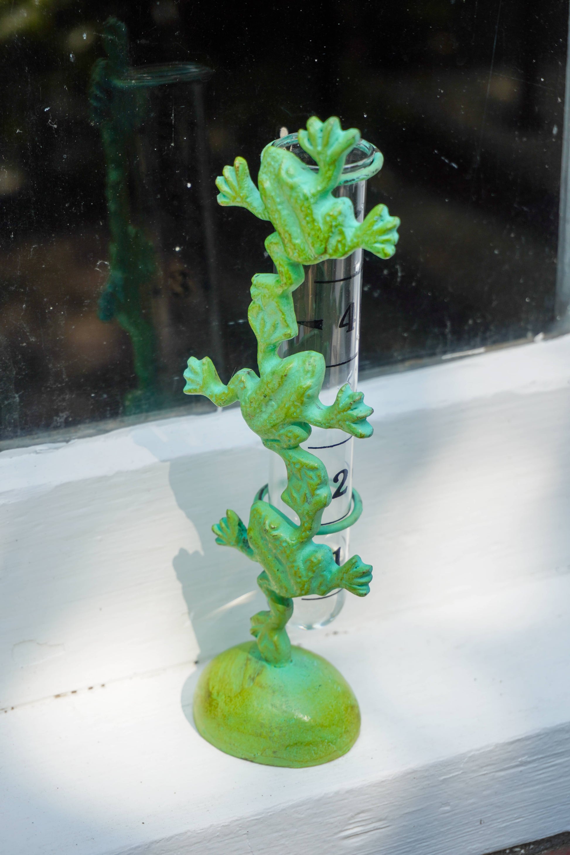 A green frog-themed thermometer decoration with three stacked frogs on a round base, shown in front of a glass window with a white painted sill.