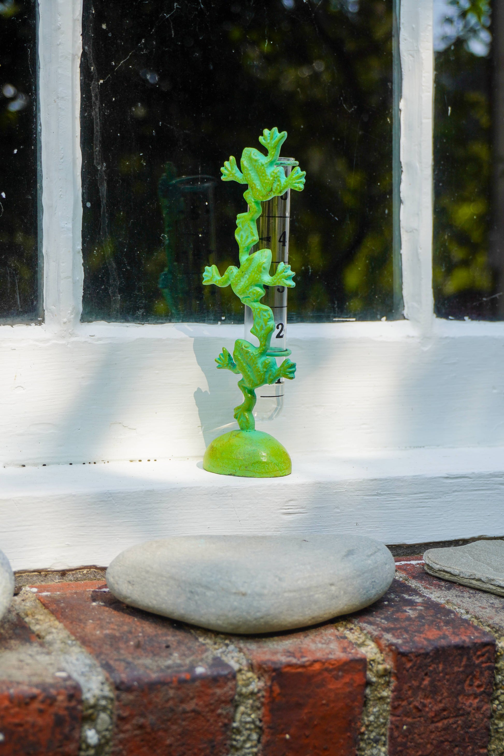 A small bright green frog-shaped decorative object standing upright on a round base, placed on a white windowsill with brick and stone visible in the foreground.