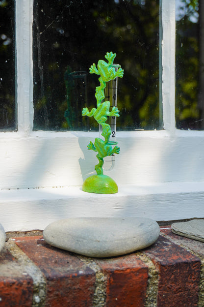 A small bright green frog-shaped decorative object standing upright on a round base, placed on a white windowsill with brick and stone visible in the foreground.