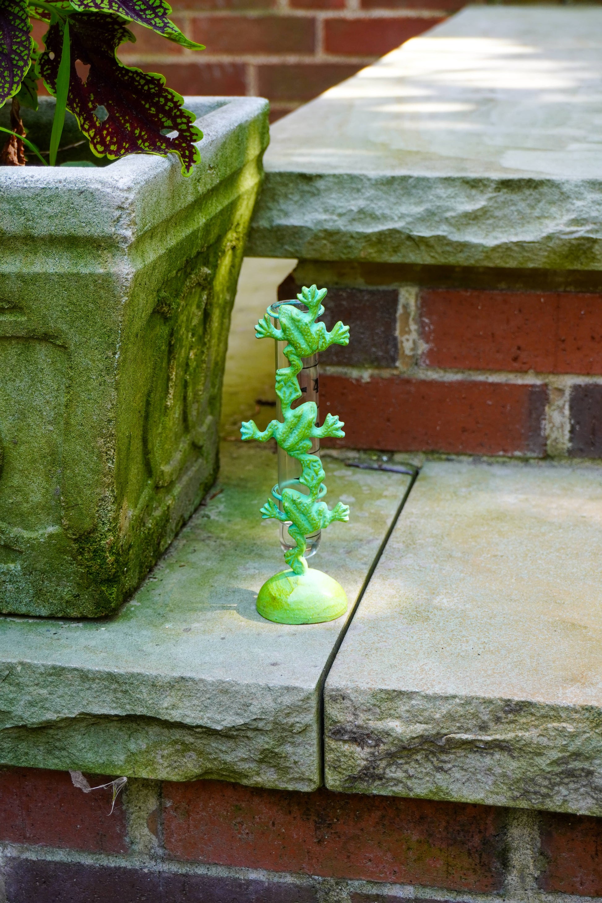 A small green frog figurine perched on a curved metal rod atop a round base, displayed on a stone ledge next to a brick wall and a stone planter.