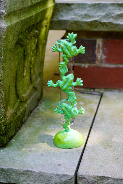 A green frog-themed rain gauge featuring a stack of three green frogs climbing up a clear glass cylinder, mounted on a round green base, set outdoors on stone pavers.