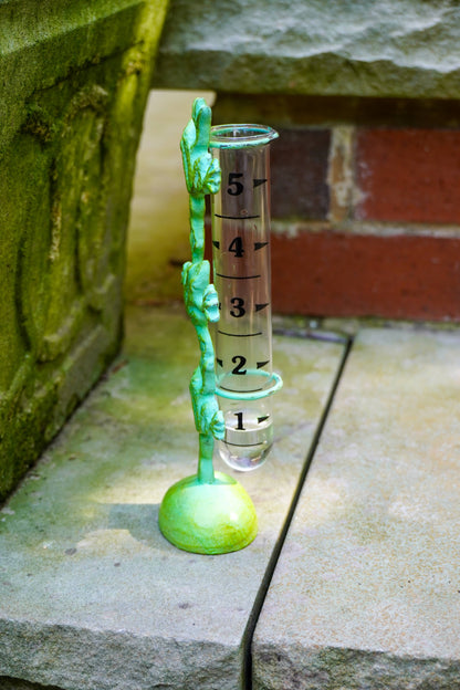 A green metal rain gauge stand with a clear graduated cylinder hanging from a decorative vine-like post, placed on a stone surface outdoors.