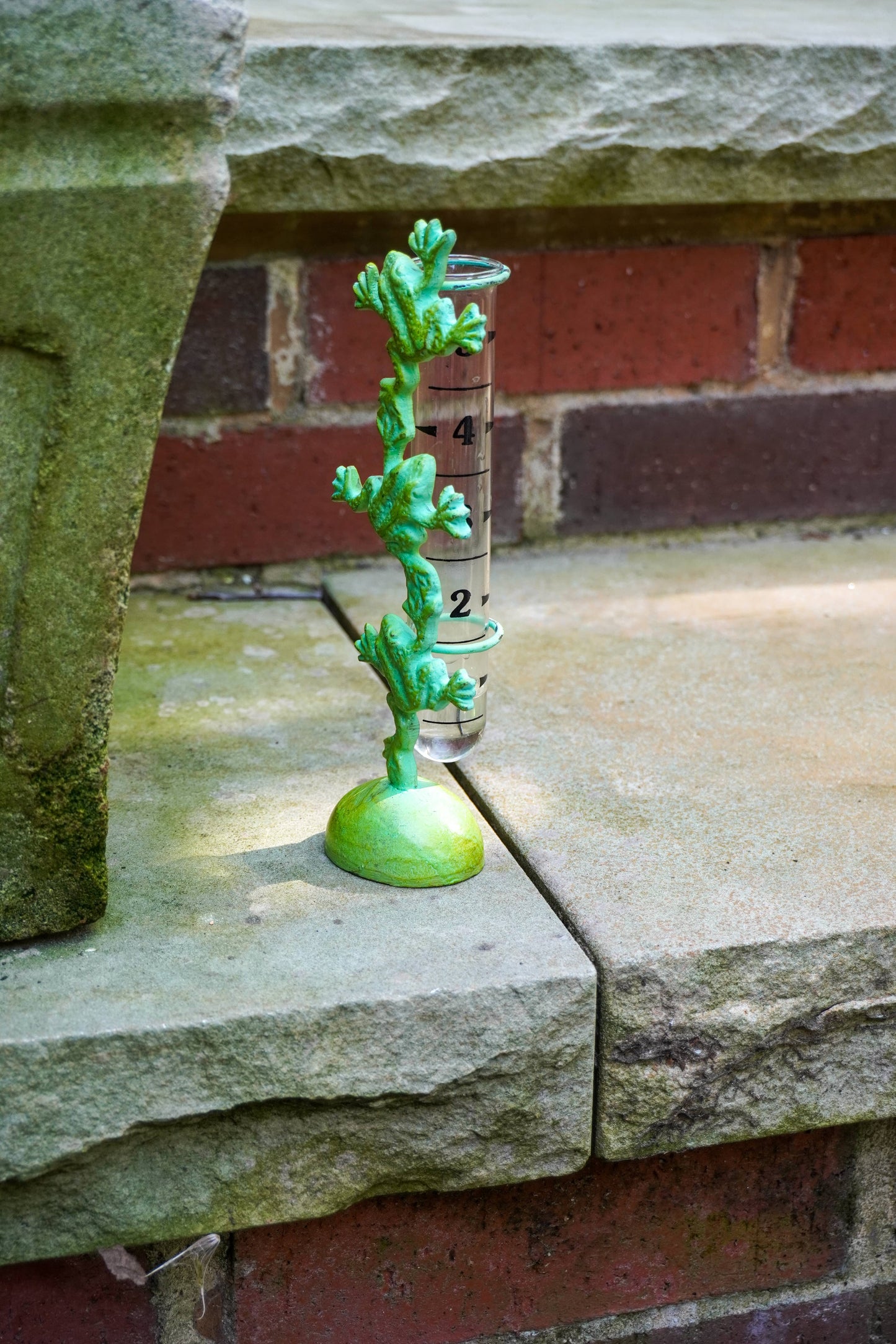 A small green plant-shaped rain gauge with a clear cylindrical tube and black measurement markings, placed on a stone ledge in front of a brick wall.