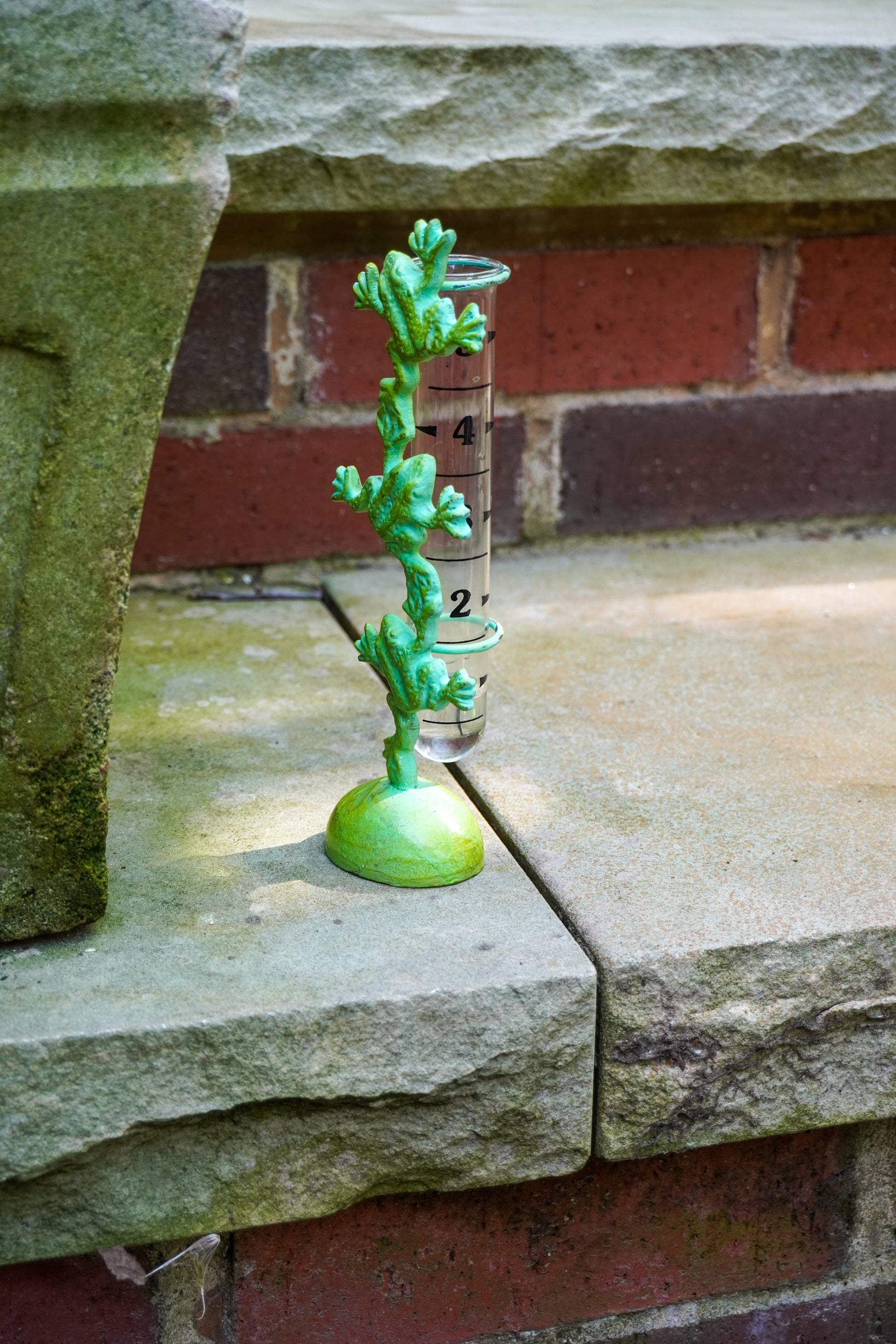 A small green plant-shaped rain gauge with a clear cylindrical tube and black measurement markings, placed on a stone ledge in front of a brick wall.