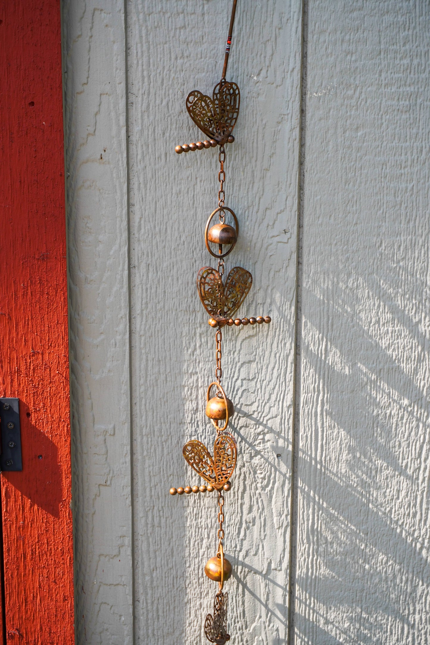 A vertical hanging metal garden ornament with ornate heart-shaped cutout pieces, small hanging bells, and chain links, photographed against a white painted wooden wall next to a red painted wooden surface.