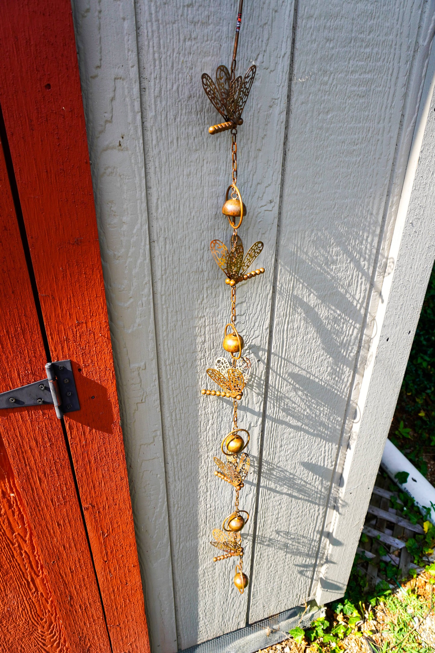A vertical hanging metal garden ornament featuring dragonfly motifs with small gold beads, photographed against a white painted wooden wall next to a red-orange door.