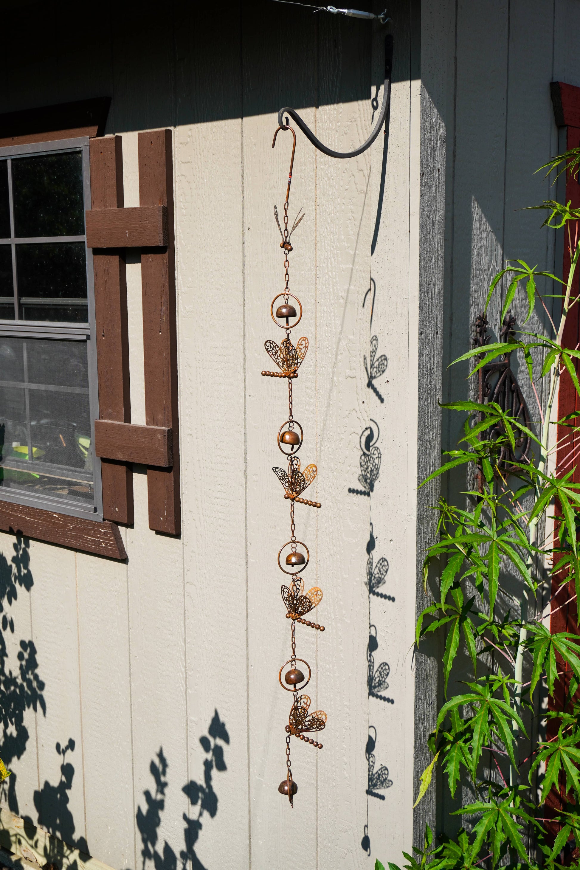 A hanging metal garden ornament with dragonfly and bell motifs, suspended outdoors against a beige house wall with green plants visible on the right.