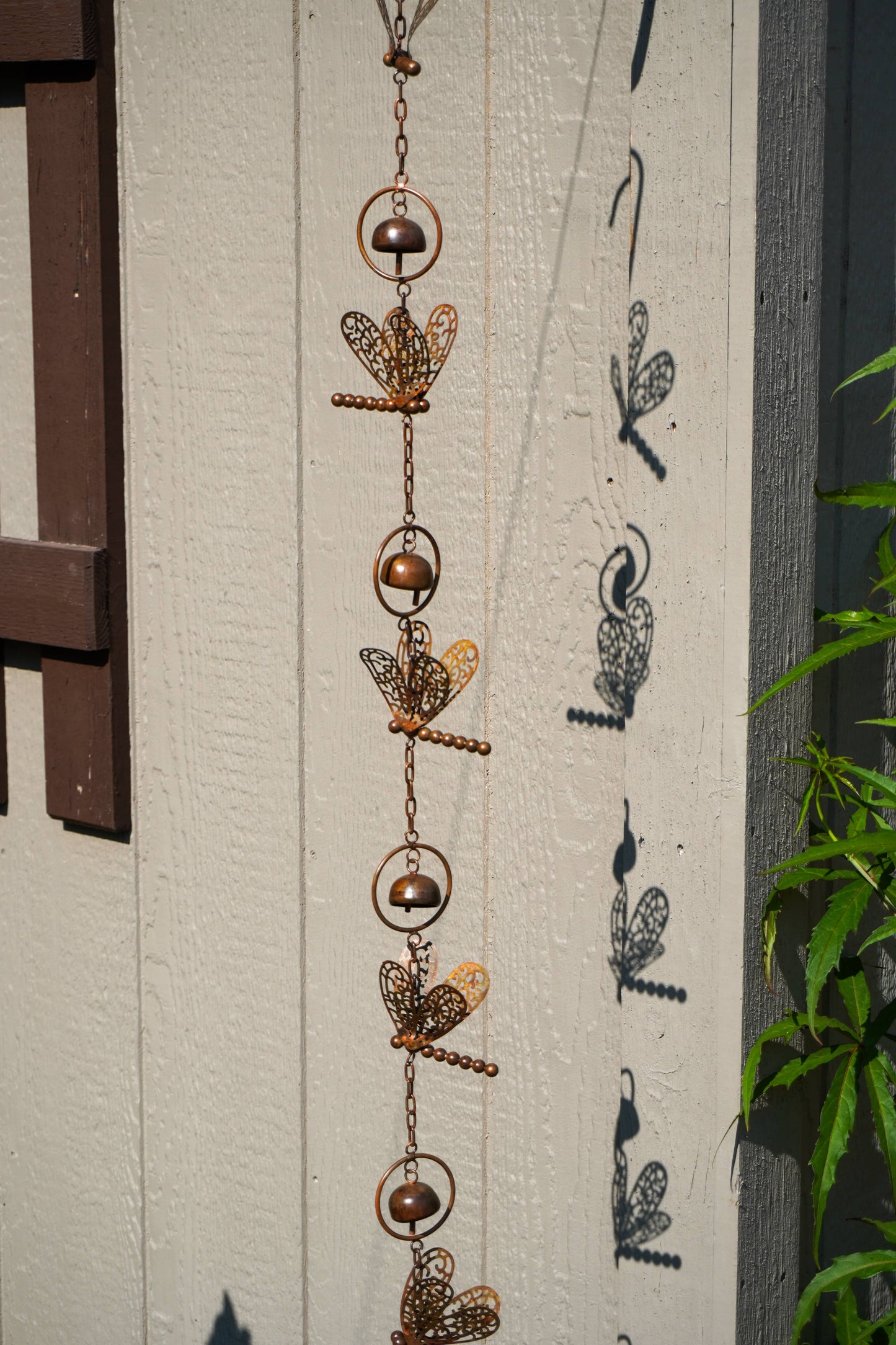 A copper-toned metal garden ornament with dragonfly designs hanging vertically against a textured off-white wall, casting shadows next to green foliage.