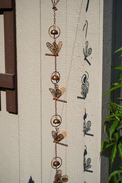 A copper-toned metal garden ornament with dragonfly designs hanging vertically against a textured off-white wall, casting shadows next to green foliage.