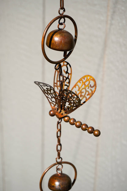 A decorative metal garden ornament featuring a dragonfly design with cutout wings, a small hanging bell, and a beaded rod, shown against a light textured wall.