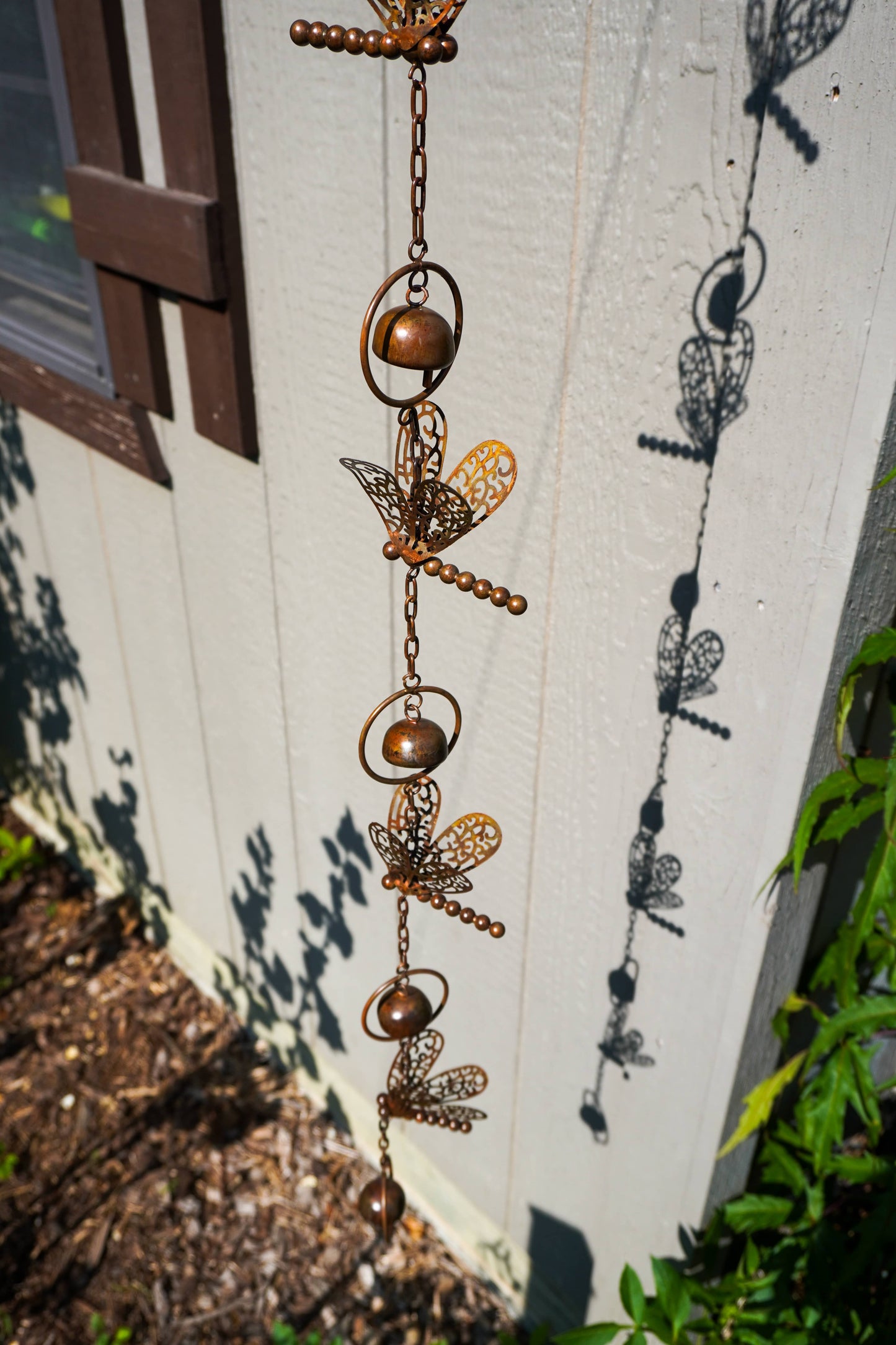 A copper-toned metal garden ornament featuring dragonfly motifs with hanging bells, shown outdoors against a white house wall with leafy green plants and a window visible.
