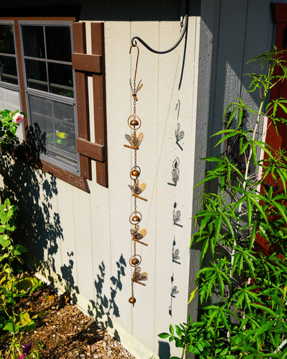 A decorative hanging garden ornament with metallic gold and bronze dragonfly and bead elements, suspended from a black wire hook on a white wooden fence in a garden setting.