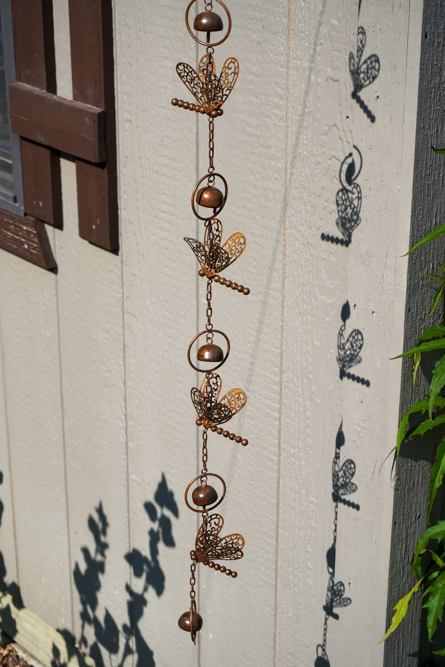 A vertical hanging metal garden ornament made of linked rings, chains, and dragonfly-shaped cutouts, shown outdoors against a white painted wooden wall with leaf shadows cast on it.