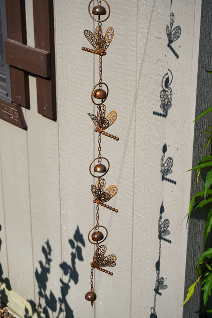 A vertical hanging metal garden ornament made of linked rings, chains, and dragonfly-shaped cutouts, shown outdoors against a white painted wooden wall with leaf shadows cast on it.