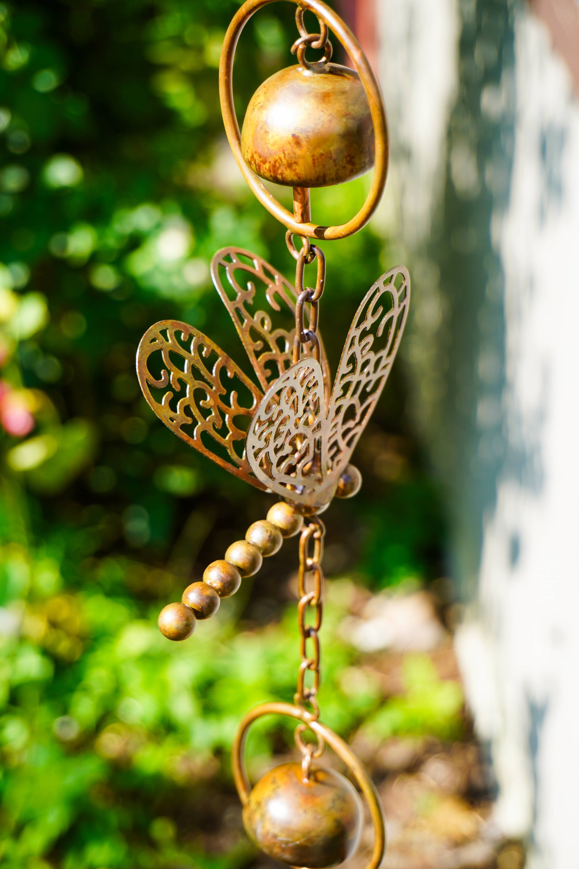 A decorative metal garden ornament featuring a dragonfly design with gold-toned beads, a small bell, and circular rings, photographed outdoors against a blurred green garden background.