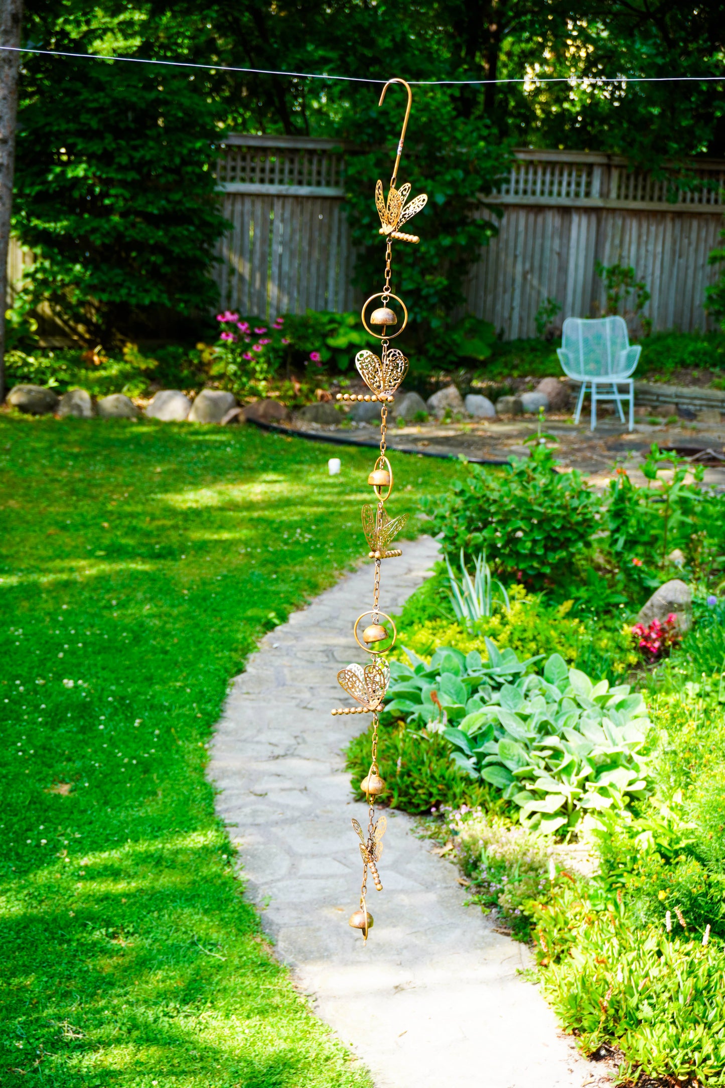 A gold metal garden wind spinner with dragonflies, and bells design hangs from a wire in a sunny backyard garden with a stone path and green grass.